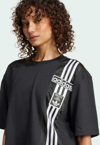 Černé oversized tričko s bílým pruhovaným designem a síťovou boční částí. Logo Adidas a detail nášivky na hrudi.