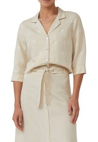 Lichtbeige button-up blouse met geborduurde vlinderpatronen, driekwart mouwen en een relaxte pasvorm. In combinatie met een bijpassende rok.