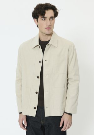 Beige lærredsjakke med knaplukning, spids krave, to forlommer og tonet syning. Båret over en mørk sweater.