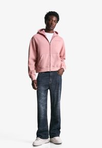 Lyserød zip-up hoodie med forlommer, båret over en hvid t-shirt. Med mørkeblå denim jeans og hvide sneakers.