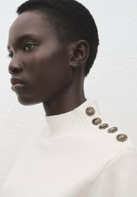 Cremefarbener Pullover mit geripptem Halsausschnitt, der sechs glänzende braune Knöpfe entlang der linken Schulter aufweist. Weiche Textur, figurbetontes Design.