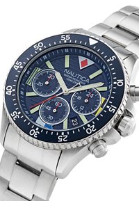 Nautica ura z nerjavečega jekla z modrim številčnikom, tremi podštevilčnicami, izrazitimi kazalci ur in teksturiranim okvirjem; vodoodporna do 100 m.