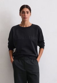 Femme avec des cheveux foncés en une basse queue de cheval, portant un sweat-shirt noir Marc O’Polo et un pantalon noir, les mains dans les poches, se tenant contre un mur blanc.