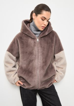 Felpa con zip in tonalità marrone e beige, dotata di cappuccio, texture morbida e vestibilità comoda. Indossata con un pullover grigio e jeans scuri.