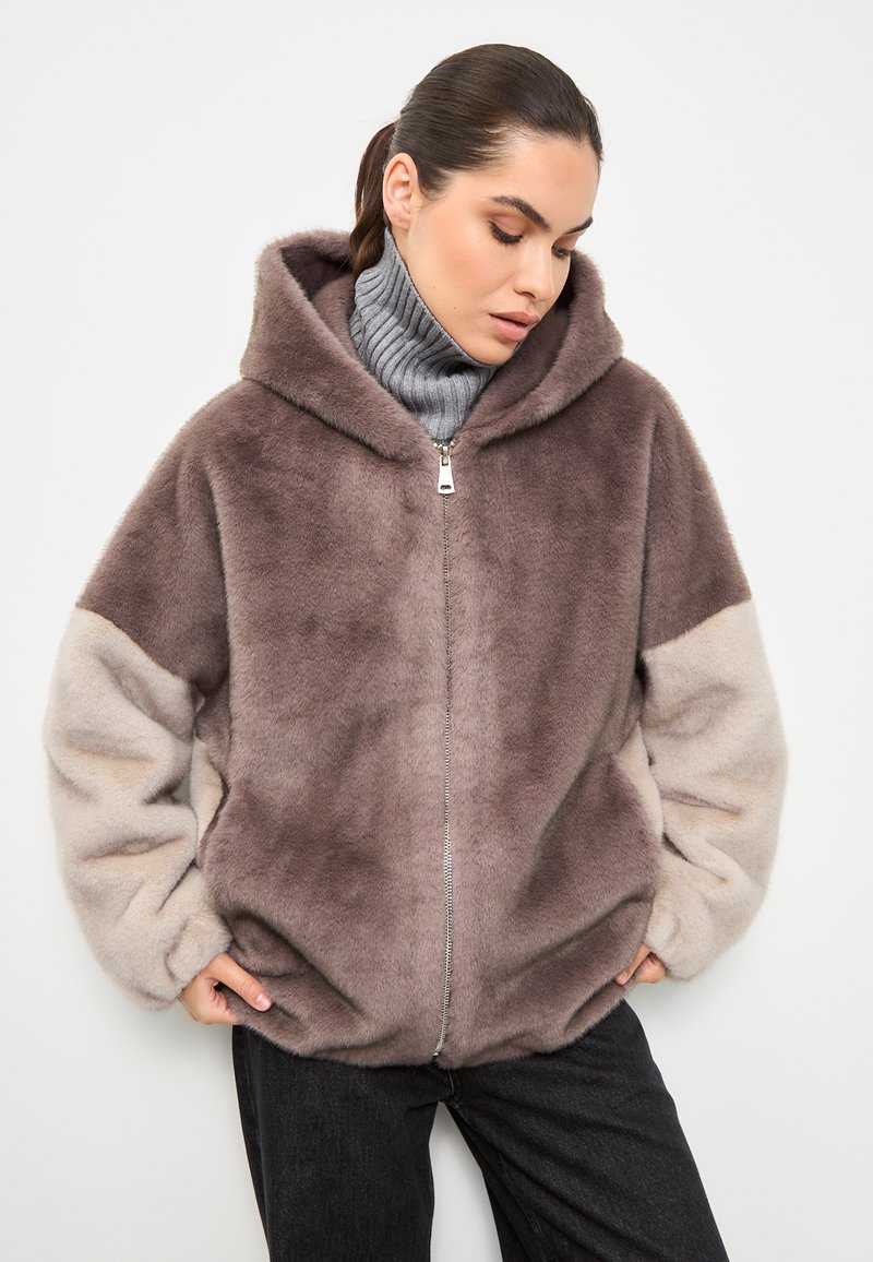 Felpa con zip in tonalità marrone e beige, dotata di cappuccio, texture morbida e vestibilità comoda. Indossata con un pullover grigio e jeans scuri.
