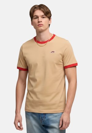 Giovane uomo che indossa una maglietta beige con colletto e polsini rossi, piccolo logo sul petto, abbinata a jeans azzurri, in piedi su sfondo bianco.