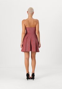 Rihmata, põlvepikkune kleit sädelevast punasest kangast, millel on liibuv korpus ja laienev seelik. Mustad kontsakingad. Tagantvaade.