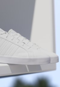 Par de zapatillas blancas de caña baja con cordones apoyadas en un borde blanco contra un fondo suave azul y gris.