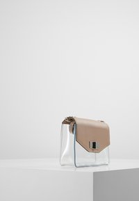 Sac à main transparent avec un rabat en cuir beige, ferrures argentées et coutures contrastées. Forme cubique avec un corps semi-transparent et un design angulaire.