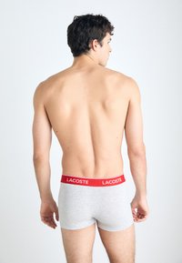 Γκρίζα σλιπάκια με κόκκινη ελαστική μέση που φέρει το λογότυπο "Lacoste". Το υλικό φαίνεται μαλακό και ελαστικό. Το μοντέλο φαίνεται από πίσω.