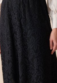 Laikkuinen tummansininen hame, jossa on kukkakuvio ja tekstuurisia yksityiskohtia, tasaisella pinnalla ja istuvalla vyötäröllä. Hieman kiiltävä pinta tuo suunnitteluun lisäilmettä.