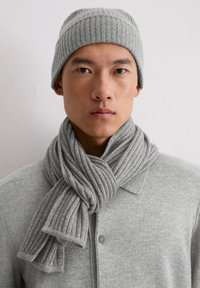 Bonnet en tricot côtelé gris et écharpe assortie, présentant une texture douce avec un design torsadé. Portés sur un cardigan gris boutonné.