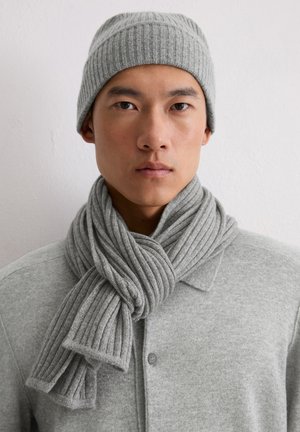 Cappello a maglia grigio a coste e sciarpa coordinata, con una texture morbida e un design intrecciato. Indossati sopra un cardigan grigio con bottoni.
