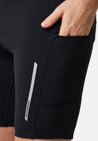 Pantaloni sportivi neri realizzati in tessuto elasticizzato, dotati di tasca laterale e striscia riflettente bianca con branding. Texture liscia e design aderente.