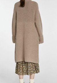 Beige neulottu neuletakki, jossa on teksturoitu kuvio, pitkät hihat ja sivuhalkiot, kerroksittain päällimmäisenä kukkakuvioisen mekon päällä, jossa on röyhelöhelma.