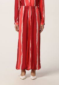 Donna che indossa pantaloni a righe verticali rossi e rosa a gamba larga con vita elasticizzata e tacchi a punta bianchi, in piedi su uno sfondo bianco.
