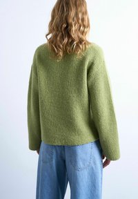 Personne aux longs cheveux ondulés portant un pull ample côtelé vert olive et un jean taille haute bleu clair, de dos sur un fond uni.
