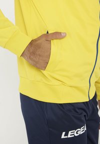 Chaqueta deportiva amarilla con cremallera y bolsillo lateral, combinada con pantalones azul marino que presentan un logo blanco. Textura suave y diseño ajustado.