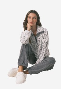 Hvid fleece-pullover med leopardprint, grå lynlås og krave, parret med grå, løstsiddende bukser og hvide strikkede hjemmesko.