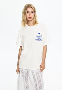 T-shirt en coton crème à coupe décontractée. Présente un motif graphique bleu et le texte "Repas Partagé" sur la zone gauche de la poitrine.