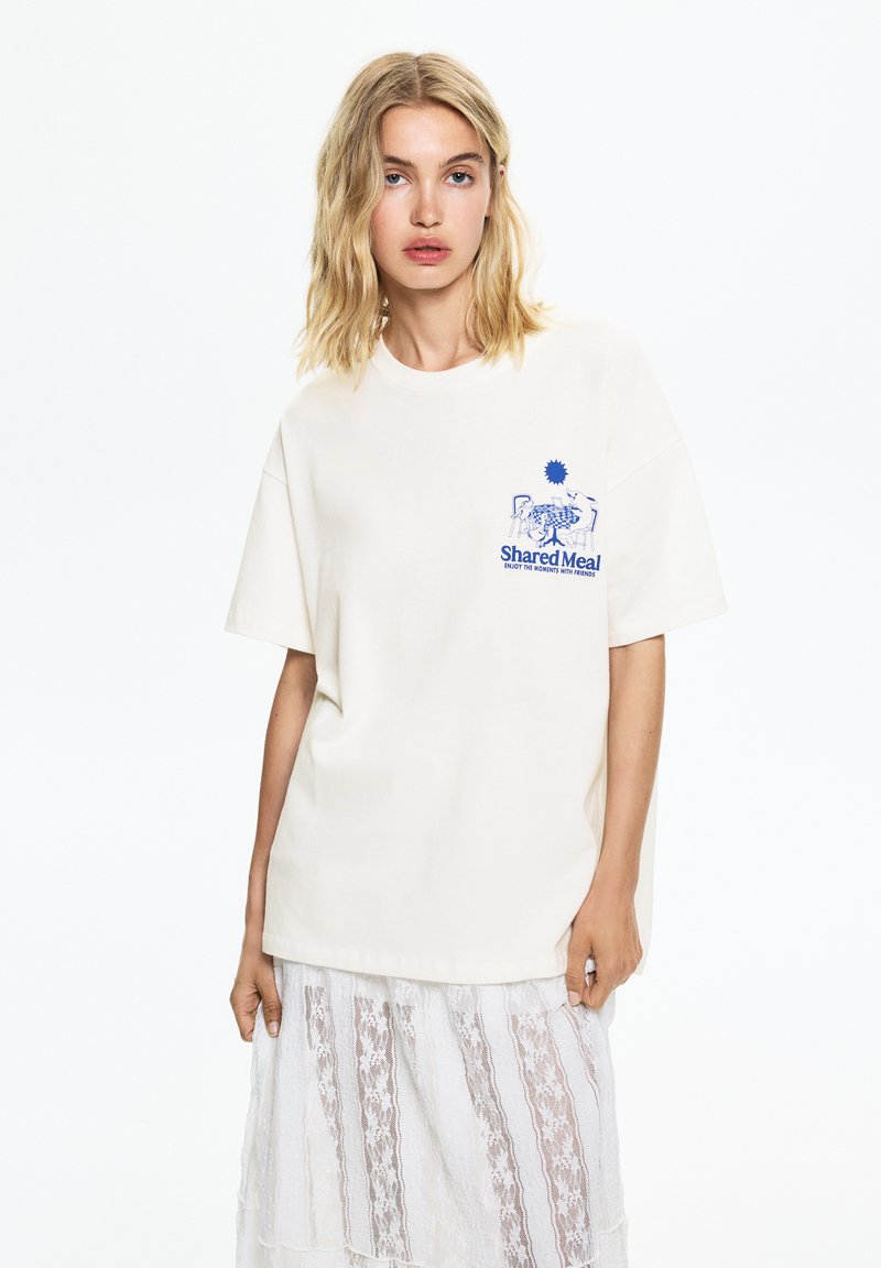 Tricou din bumbac crem, cu o croială lejeră. Prezintă un design grafic albastru și textul "Masă Comună" în zona stângă a pieptului.
