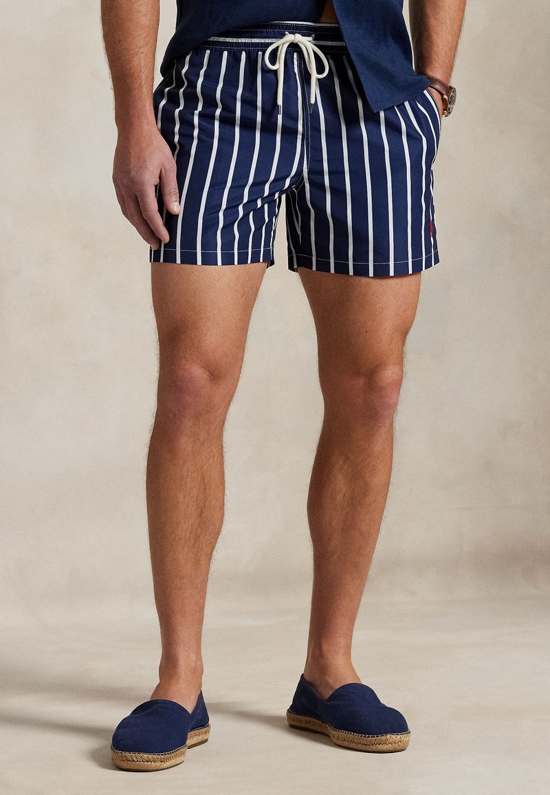 Calções de banho navy com riscas verticais brancas, cintura elástica e cordão, combinados com espadrilhas azuis. Tecido texturizado.