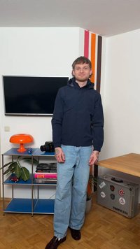 Άνδρας με τατουάζ στα χέρια, φορώντας μπλε σακάκι και ανοιχτόχρωμο τζιν, στέκεται σε ένα δωμάτιο με τηλεόραση, πορτοκαλί λάμπα, βιβλιοθήκη και βαλίτσα.