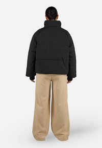 Svart pufferjakke med høy krage, vid silhuett og ribbestrikkede mansjetter. Bært med lysebeige, vide bukser, sett fra baksiden.