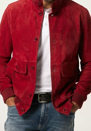 Mies, jolla on punainen avonainen mokkatakki valkoisen paidan päällä, kädet sinisten farkkujen taskuissa ja näkyvillä musta vyö.