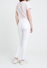 Mujer vistiendo una camiseta rosa clara, pantalones vaqueros skinny blancos y zapatillas Reebok blancas, de pie contra un fondo blanco liso.