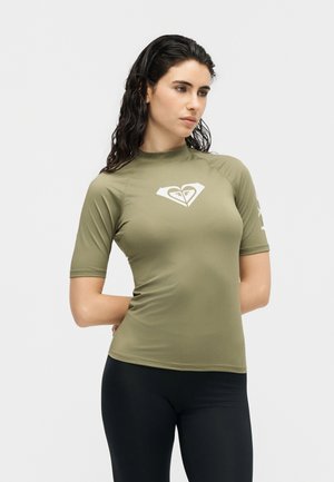 Žena s mokrom kosom koja nosi maslinasto zelenu kratku rash guard majicu s bijelim logotipom u obliku srca, uz crne tajice, stoji na bijeloj pozadini.