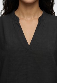 Femme portant un haut noir en tissu texturé avec un décolleté en V et un petit col montant.