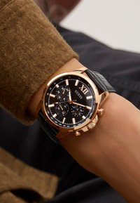 Montre dorée avec un cadran noir, fonction chronographe, et un bracelet en cuir noir texturé ; index horaires blancs et un grand chiffre romain XII.