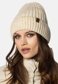 Beige geribbelde gebreide muts met een omgeslagen rand en een klein leren label aan de zijkant, gedragen door een vrouw in een lichtgekleurde jas.
