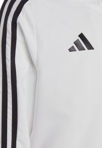 Witte sportjacket met zwarte strepen op de mouwen en een klein zwart logo. De stof lijkt glad met een lichte glans.