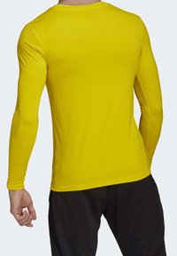 Man som bär en klar gul långärmad skjorta och svarta byxor, sedd bakifrån mot en enkel ljus bakgrund.