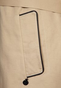 Beige stoffen zak met zwarte rand, fijne stiksels en een klein metalen accent. De zak is rechthoekig met strakke randen.