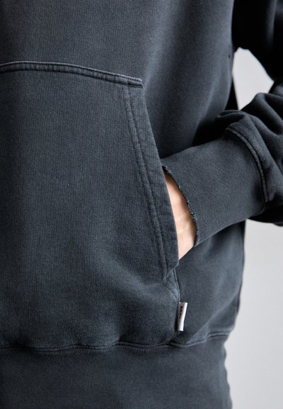 Sudadera gris oscuro con un bolsillo frontal de borde sin rematar, puños acanalados y una pequeña etiqueta en la costura lateral. Tejido pesado con una textura suave.