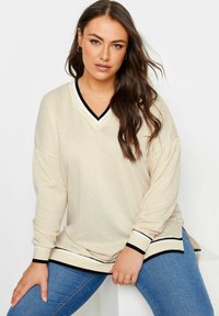 Maglione oversize beige con scollo a V, motivo testurizzato, bordo nero e crema sul colletto e sui polsini, maniche lunghe e spacchi laterali.