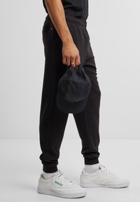 Pantalones de chándal negros con cintura elástica, bolsillo lateral y puños acanalados. Combinados con zapatillas blancas y una gorra negra sostenida en la mano.