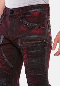 Rote und schwarze Denim-Hosen mit strukturierten Einsätzen, Reißverschlüssen und dekorativen Nieten, die ein enges Design und distressed Akzente aufweisen.