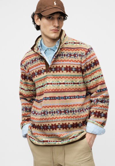 Noor mees pruun mütsiga, helebeeži mustriga fliisist pullover üle helesinise kraega särgi ja beežide pükstega, käed taskutes, neutraalne taust.