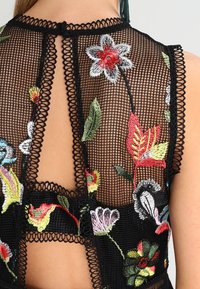 Czarny siateczkowy top z kolorowym haftem kwiatowym. Z tyłu zapinanie na guziki w kształcie łezki, z falbaniastymi brzegami oraz skomplikowanymi detalami haftu.