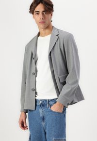 Sivša blazer s sproščenim krojem, dvema gumboma, koničastimi lapelami in enim žepom na prsih. Nosi se preko belega majice in modrih kavbojk.