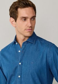 Modrá riflová košela s golierom na gombíky, bielymi gombíkmi a vyšitým logom na prsiach. Hladká textura a uvoľnený strih.