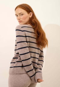 Beżowy sweter w niebiesko-beżowe paski o miękkiej fakturze, z okrągłym dekoltem i ściągaczami. Noszony z dopasowanymi jasnobeżowymi dołami.