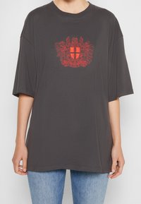 Tmavě šedé oversized tričko s krátkými rukávy, na přední straně zdobené vyšitým erbem červené barvy. Hladká textura, ležérní střih.