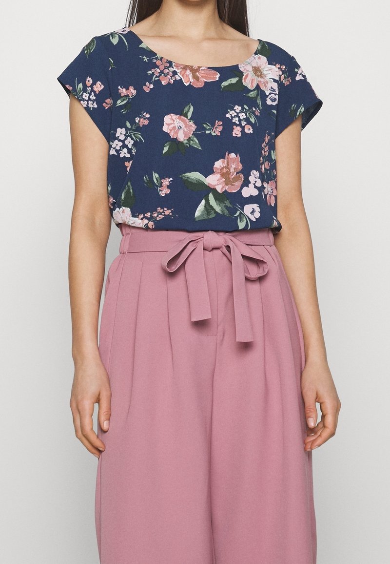 Femme portant un haut à manches courtes bleu marine à motif floral, rentré dans un pantalon taille haute rose poussiéreux à jambes larges, ceinturé d'un ruban en tissu noué à l'avant.