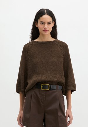 Vrouw met lang donker haar draagt een losse bruine gebreide trui en bruine short met hoge taille, een zwarte riem en een gouden gesp.