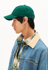 Junger Mann mit grünem Baseballcap, geschichteter grüner und beiger karierten Hemd und blauer Jeansjacke mit beigem Kragen, der links vor einem weißen Hintergrund schaut.